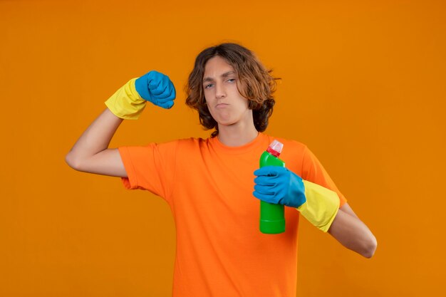 Joven en camiseta naranja con guantes de goma sosteniendo una botella de artículos de limpieza apretando el puño regocijándose de su éxito y victoria mirando confiado, satisfecho y orgulloso de pie ov