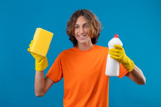 Joven en camiseta naranja con guantes de goma sosteniendo una botella de productos de limpieza y una esponja mirando a la cámara sonriendo alegremente de pie sobre fondo azul.