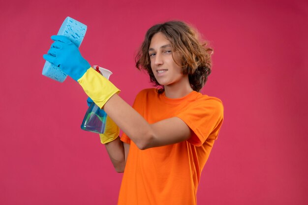 Joven en camiseta naranja con guantes de goma sosteniendo spray de limpieza y una esponja mirando confiado sonriendo de pie sobre fondo rosa