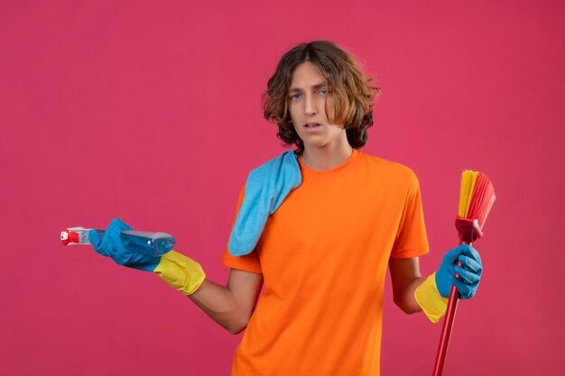 Joven en camiseta naranja con guantes de goma sosteniendo un trapeador y spray de limpieza mirando confundido de pie sobre fondo rosa