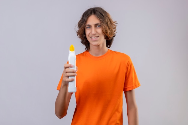 Joven en camiseta naranja sosteniendo una botella de productos de limpieza mirando a la cámara con una sonrisa escéptica de pie sobre fondo blanco.