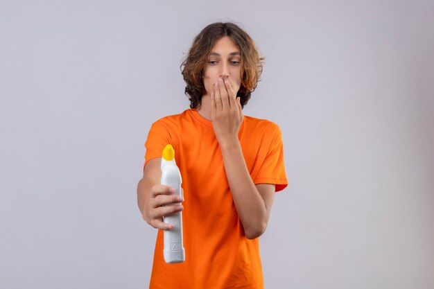 Joven en camiseta naranja sosteniendo una botella de productos de limpieza mirándolo sorprendido y asombrado cubriendo la boca con la mano de pie sobre fondo blanco.