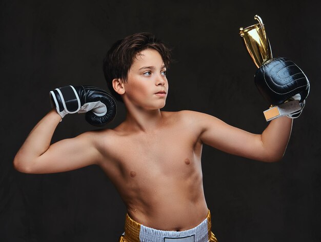 El joven campeón de boxeador sin camisa que usa guantes sostiene una copa de ganador que muestra los músculos. Aislado en un fondo oscuro.