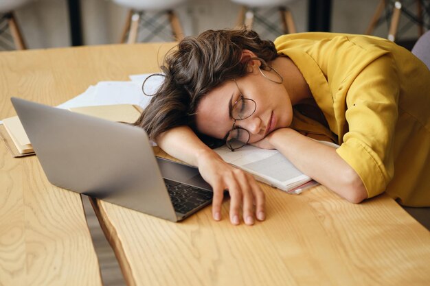 Una joven cansada con anteojos se queda dormida en el escritorio con una computadora portátil y documentos debajo de la cabeza en el lugar de trabajo