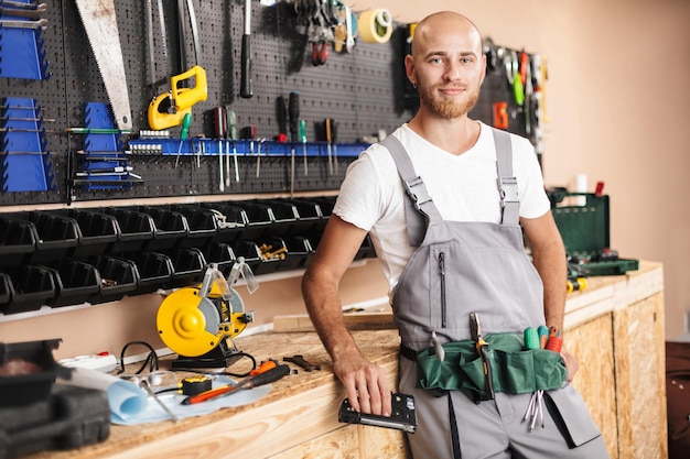 Foto gratuita joven capataz sonriente en ropa de trabajo sosteniendo una grapadora en la mano mirando soñadoramente a la cámara con soporte de diferentes herramientas en el fondo del taller