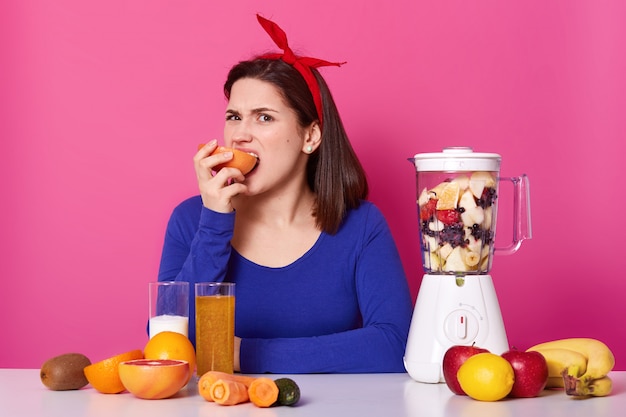 Joven carismática, lleva una diadema roja brillante y un suéter azul, muerde y come toronja, no sabe bien, licuado de frutas en la licuadora, mucha comida saludable frente a la señora que come. Dieta saludable.