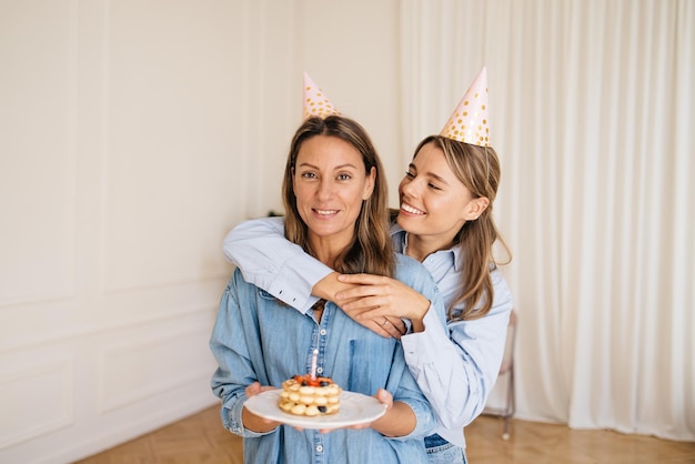 Una joven caucásica abraza a su madre desde atrás sosteniendo un pastel con una vela en la sala de luz. Concepto de cumpleaños, dulces amorosos.