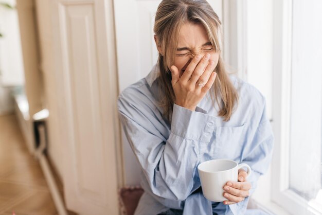 Una joven caucásica alegre bebe café y se ríe mientras se cubre la cara con la mano mientras se sienta junto a la ventana en la sala de luz. Concepto de emoción