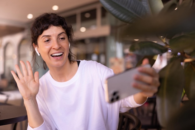 Una joven caucásica alegre con el pelo recogido oscuro en una blusa blanca saludando a la cámara de la pantalla del teléfono en el café