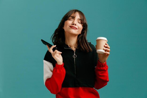 Una joven caucásica complacida sosteniendo un teléfono móvil y una taza de café para llevar con los ojos cerrados aislada de fondo azul