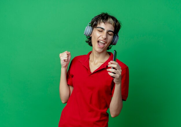 Joven caucásica con corte de pelo pixie usando audífonos escuchando música sosteniendo el teléfono móvil finge cantar usando el teléfono como micrófono con el puño cerrado y los ojos cerrados