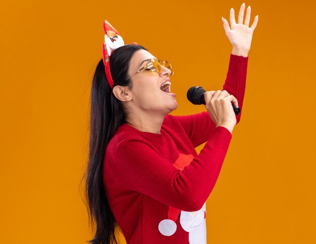 Joven caucásica con diadema de santa claus y suéter con gafas de pie en la vista de perfil sosteniendo el micrófono cerca de la boca cantando con los ojos cerrados levantando la mano aislada en la pared naranja con espacio de copia