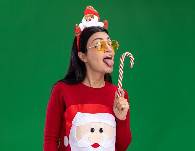 Joven caucásica con diadema de santa claus y suéter con gafas sosteniendo y mirando el tradicional bastón de caramelo navideño mostrando la lengua preparándose para lamerlo aislado en la pared verde con espacio de copia