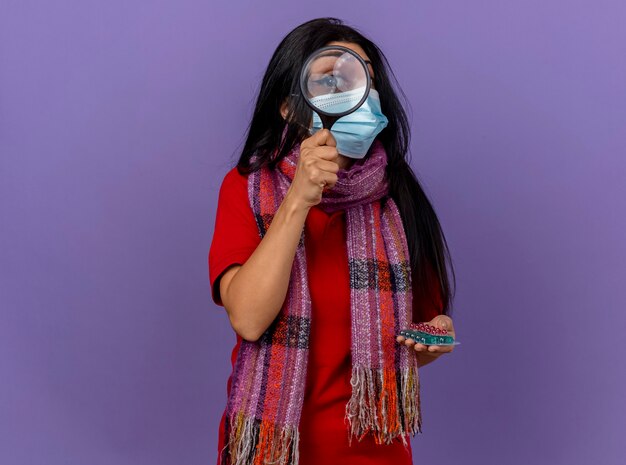 Joven caucásica enferma con máscara y bufanda sosteniendo paquetes de cápsulas a través de una lupa aislada en la pared púrpura con espacio de copia