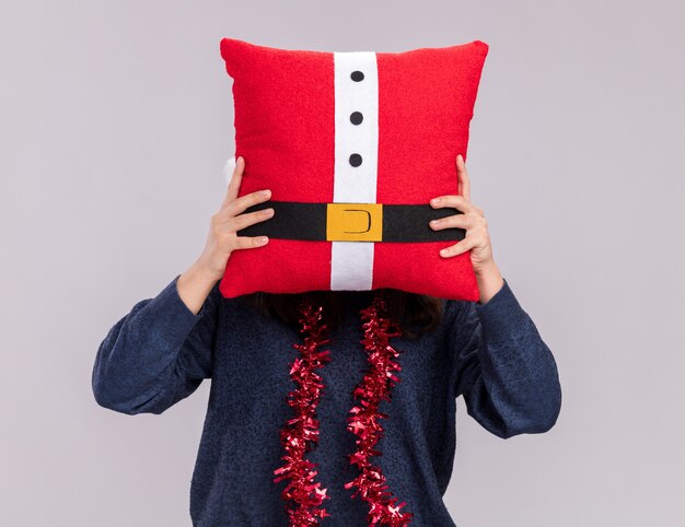 Joven caucásica con gorro de Papá Noel y guirnalda alrededor del cuello sostiene una almohada decorada delante de su cabeza aislada sobre fondo blanco con espacio de copia