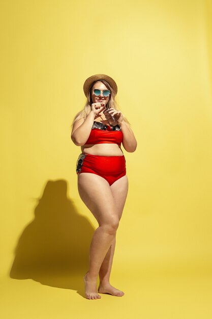 Joven caucásica modelo femenino de talla grande preparándose para resort de playa en la pared amarilla. Mujer en traje de baño rojo, sombrero y gafas de sol. Concepto de verano, fiesta, cuerpo positivo. Beber cóctel.