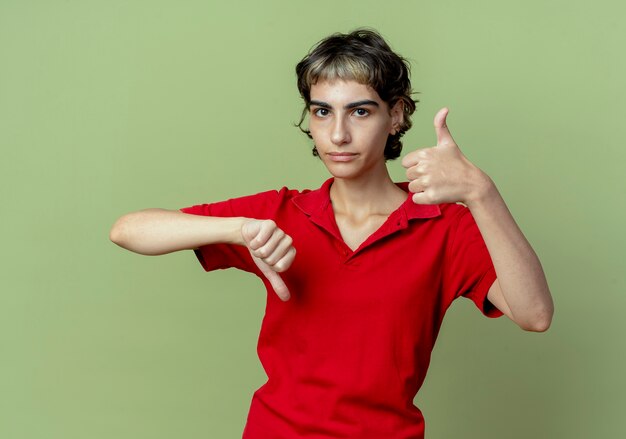 Joven caucásica molesta con corte de pelo pixie mostrando los pulgares hacia arriba y hacia abajo aislado sobre fondo verde oliva