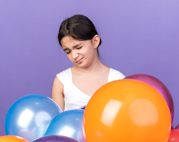 Joven caucásica molesta sosteniendo globos de helio aislado en la pared púrpura con espacio de copia