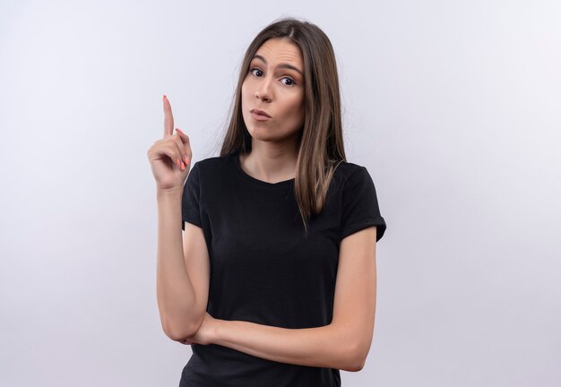 Joven caucásica vestida con camiseta negra apunta hacia arriba con el dedo en la pared blanca aislada