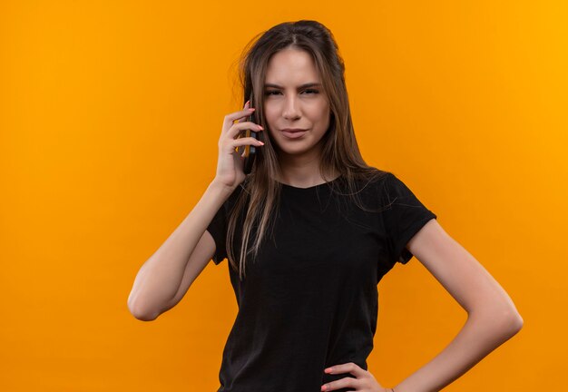 Joven caucásica vestida con camiseta negra habla por teléfono con la mano en la cadera en la pared naranja aislada