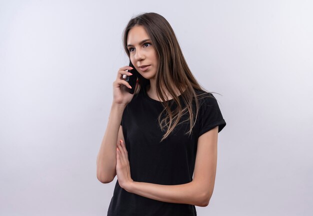 Joven caucásica vestida con camiseta negra habla por teléfono en la pared blanca aislada