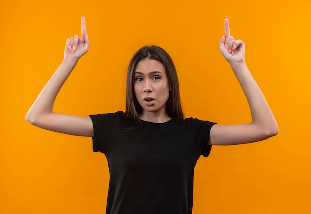 Joven caucásica vistiendo camiseta negra apunta hacia arriba con ambas manos en la pared naranja aislada