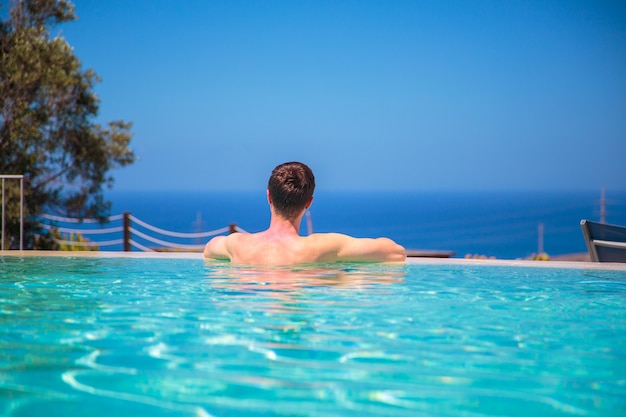 Joven caucásico en la piscina infinita mirando la vista al mar, relajándose y disfrutando de su vida