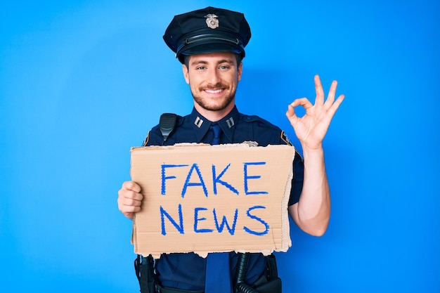 Foto gratuita joven caucásico con uniforme de policía sosteniendo una pancarta de noticias falsas haciendo un buen signo con los dedos, sonriendo amigablemente haciendo un gesto excelente