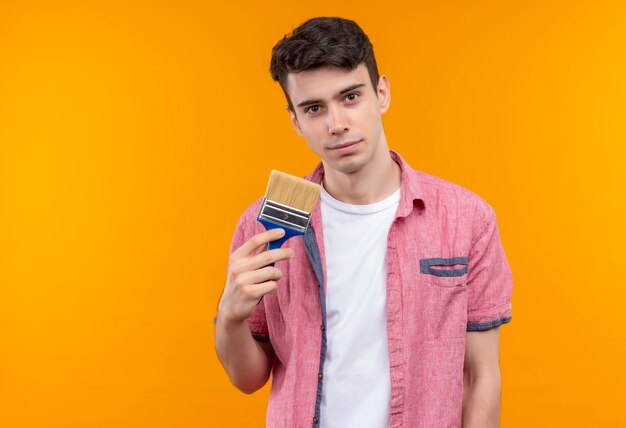 Joven caucásico vistiendo camisa rosa sosteniendo pincel sobre pared naranja aislada