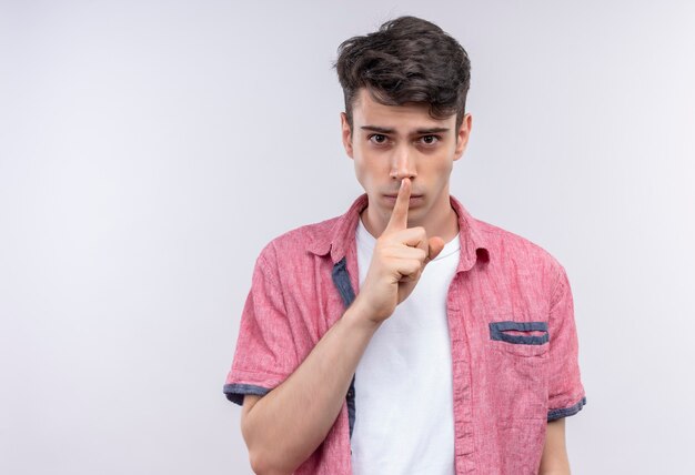 Joven caucásico vistiendo camiseta rosa mostrando gesto de silencio en la pared blanca aislada