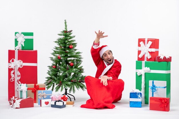 Joven celebra la fiesta de Navidad sentado en el suelo mostrando algo cerca de regalos y árbol de Navidad decorado