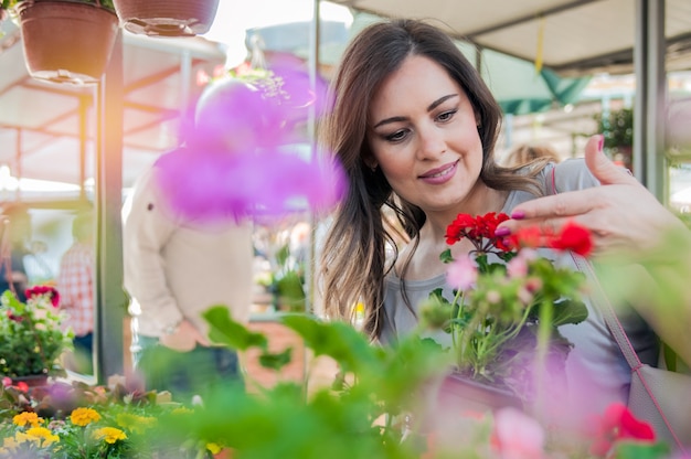 Joven celebración de geranio en olla de barro en el centro de jardinería. Flores de compras de mujer joven en el centro de jardín de mercado