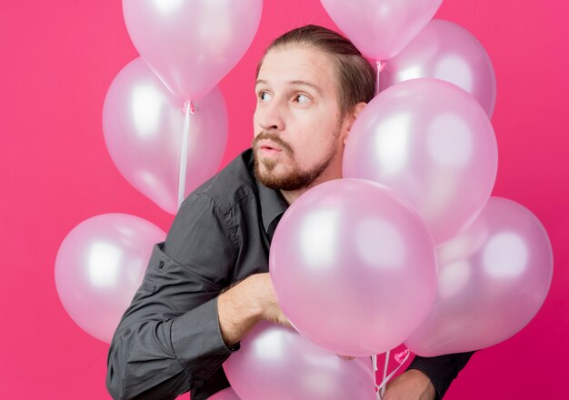 Joven celebrando la fiesta de cumpleaños con un montón de globos mirando a un lado sorprendido parado sobre la pared rosa