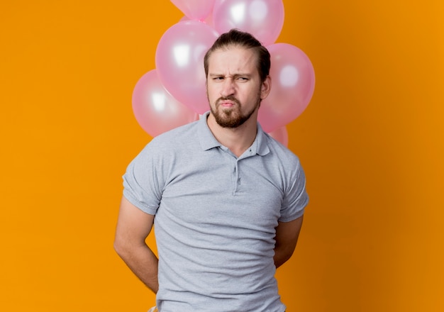 Joven celebrando la fiesta de cumpleaños sosteniendo un montón de globos mirando a un lado disgustado e infeliz de pie sobre la pared naranja