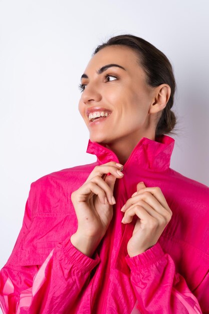 Una joven con una chaqueta deportiva rosa brillante de fondo blanco sonríe alegremente con un marco vertical