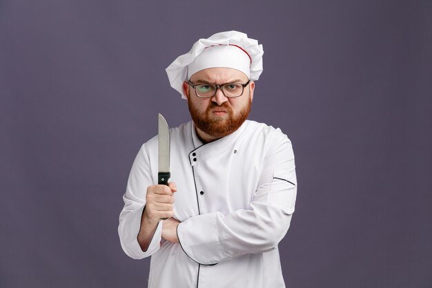 Joven chef agresivo con uniforme de anteojos y gorra sosteniendo un cuchillo mirando a la cámara mientras mantiene la mano bajo el brazo aislada en el fondo morado