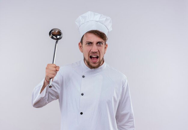 Un joven chef barbudo agresivo en uniforme blanco sosteniendo la cuchara mientras mira en una pared blanca