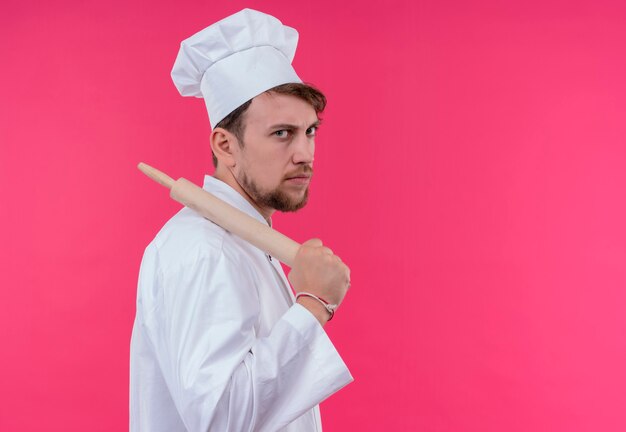 Un joven chef barbudo agresivo con uniforme blanco sosteniendo un rodillo mientras mira en una pared rosa