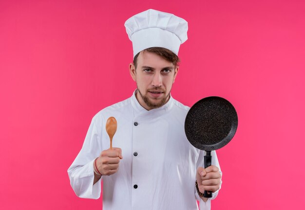 Un joven chef barbudo agresivo en uniforme blanco sosteniendo una sartén con cuchara de madera mientras mira en una pared rosa