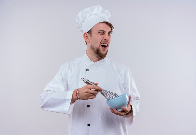 Un joven chef barbudo agresivo vestido con uniforme de cocina blanco y sombrero sosteniendo una cuchara mezcladora en un tazón azul mientras mira en una pared blanca