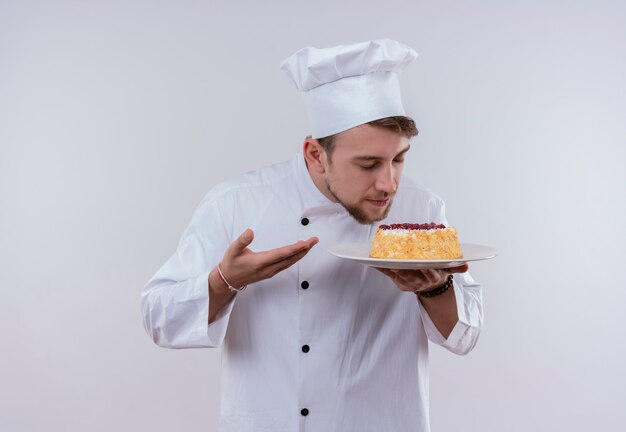 Un joven chef barbudo complacido con uniforme blanco de cocina y sombrero sosteniendo un plato con pastel y oliéndolo mientras está de pie sobre una pared blanca