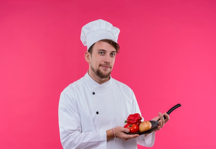 Foto gratuito un joven chef barbudo complacido en uniforme blanco sosteniendo una sartén con verduras frescas como cebolla, tomate y pimiento mientras mira una pared rosa