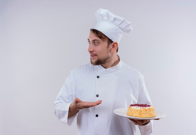 Un joven chef barbudo complacido con uniforme de cocina blanco y sombrero sosteniendo un plato con pastel mientras mira de lado en una pared blanca