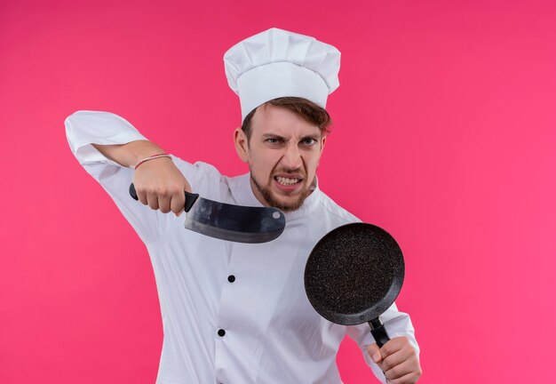 Un joven chef barbudo enojado con uniforme blanco sosteniendo un cuchillo de carnicero con una sartén mientras mira en una pared rosa