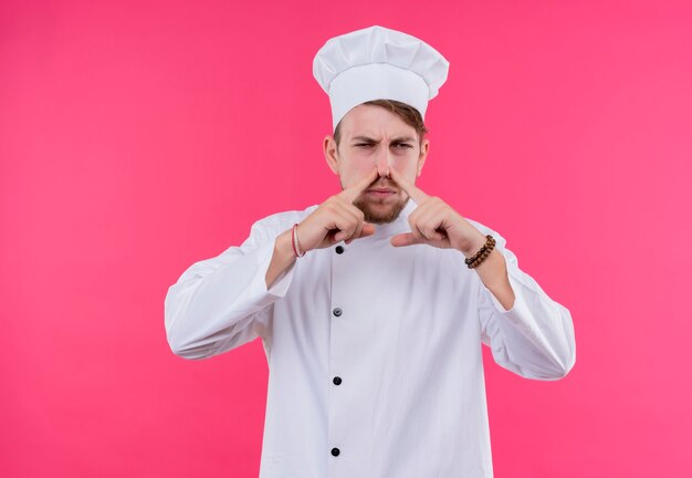 Un joven chef barbudo enojado con uniforme blanco sosteniendo los dedos índices en la nariz en una pared rosa