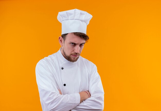 Un joven chef barbudo enojado en uniforme blanco tomados de la mano cruzados mientras mira por encima de la pared naranja