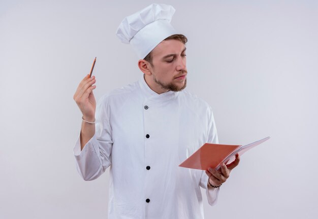 Un joven chef barbudo hombre vestido con uniforme de cocina blanco y sombrero mirando portátil y tomando notas en una pared blanca