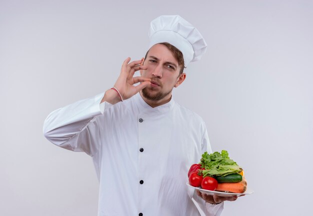 Un joven chef barbudo hombre vestido con uniforme de cocina blanco y sombrero mostrando sabroso gesto ok mientras sostiene un plato blanco con verduras frescas como tomates, pepinos, lechugas y mirando un