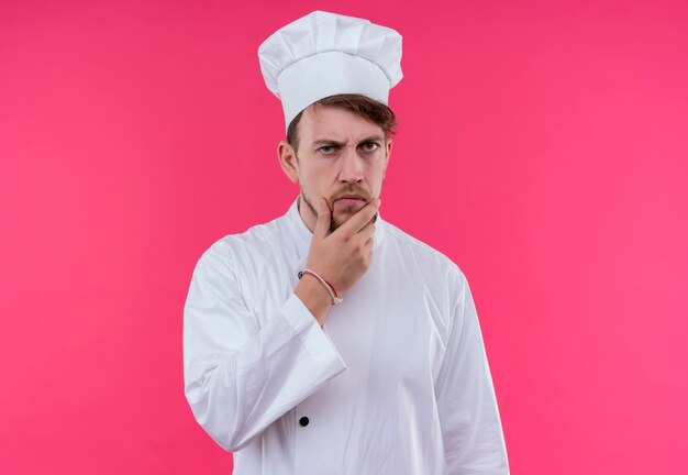 Un joven chef barbudo pensativo en uniforme blanco pensando con la mano en la barbilla mientras mira en una pared rosa