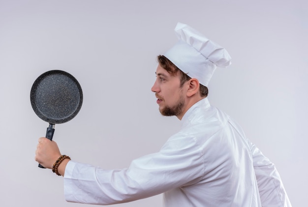 Un joven chef barbudo positivo hombre vestido con uniforme de cocina blanco y sombrero sosteniendo una sartén como un bate de béisbol en una pared blanca